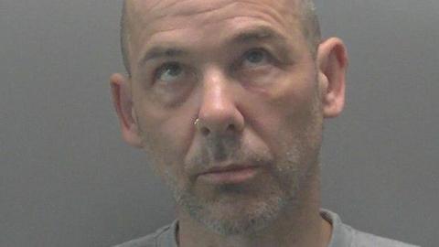 A custody image of Gavin Bainbridge - a man with a shaved head and a grey stubble beard. His eyes are looking up towards the ceiling.