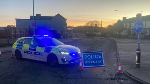 It's dusk and a police car with lights on is blocking a road. There's a sign deterring people from entering. Houses can be seen in background on the side of the road.