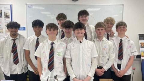 Ten schoolboys are standing in a classroom wearing white shirts that have been signed with colourful pens. Seven of them are wearing maroon and navy striped ties. There is a wall-mounted whiteboard in the background.