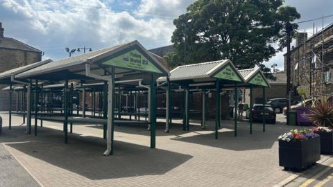 Empty market stalls painted green in Pudsey Market Square on a sunny day.