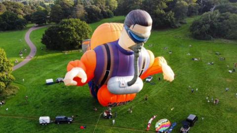 A large orange and purple Action Man hot air balloon is inflated in a grassy area surrounded by spectators and trees.
