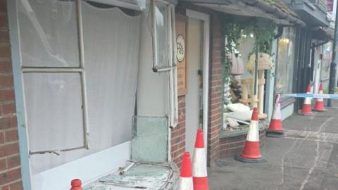 A broken shop window with shards of glass. There are traffic cones outside the properties and police tape to the right. 