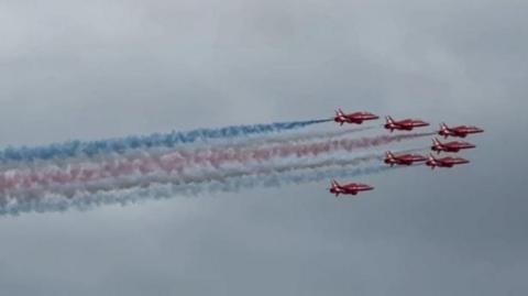 The Red Arrows in formation with white, blue and red smoke coming from their tails.