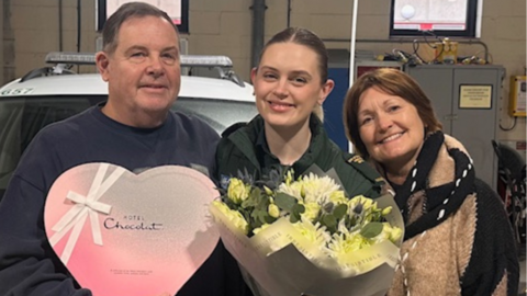 Simon Thompson is on the left and has short brown hair. He is wearing a navy jumper and is holding a heart shaped box of chocolates. Chloe Spencer is in the middle and has her hair in a ponytail. She is holding a bunch of flowers. Tina Thompson is on the right and has short brown hair and is wearing a black and beige coat.