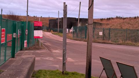 The entrance to the landfill site is surrounded by green fences with red and white signs attached. 