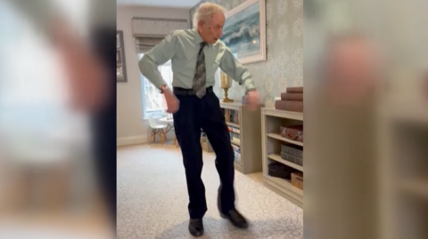 A man with grey hair, black trousers and a smart shirt, dancing