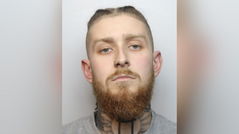 A mugshot of Alfie Wheatley, a 21-year-old wearing a grey sweater, with tattoos on his neck and a beard, wearing a nose ring and hair tied back.