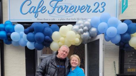 A man and a woman stood in front of a cafe on its opening day, smiling at the camera.
