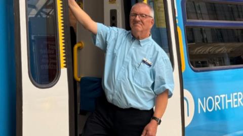 A colour image of Mr Clarke stood in the doorway of a train carriage. He is wearing spectacles and a uniform, comprising of a blue shirt and black trousers.