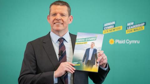 Rhun ap Iorwerth stood in front of a green wall that promotes Plaid Cymru and holds a manifesto