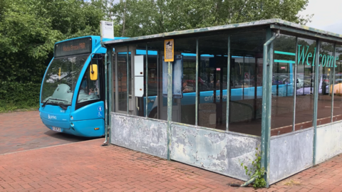 A large, enclosed bus shelter, with sheets of metal at the bottom, with a blue single-decker bus visible behind it. Trees can be seen in the background.