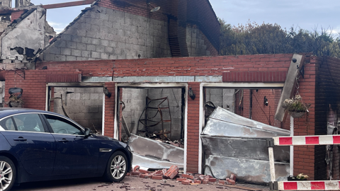 A blue car in the foreground on the left is parked in front of the remains of a garage. The adjoining house has been destroyed by a fare, with the roof missing and breeze blocks exposed