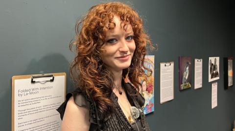 Sinead Diamond standing next to her artwork at Tullie. She has red curly hair down to her shoulders and a fringe. She has blue eyes and is wearing a black blouse. She has her back to a grey wall with artwork displayed and she is looking at the camera and smiling