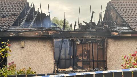 A house with part of the roof burnt out. A fence and police tape are to the forefront.