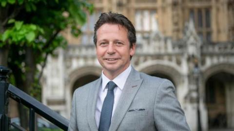 Nigel Huddlestone MP in a grey suit, white shirt and dark grey tie standing outside the Houses of Parliament