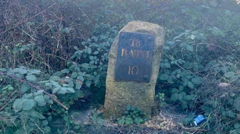 An 18th century milestone on the side of a road surrounded by overgrown shrubs. The milestone reads "To BATH, 10" and appears to have a chunk missing from its right corner (left from viewers perspective).