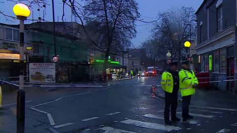 Two police officers stand on a zebra crossing with their backs facing a cordon at a murder investigation scene on a road lined with shops in Bristol. It is a grey and damp morning, which is still quite dark. A police federation van is parked beside them.