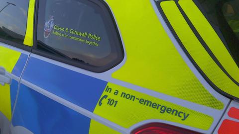 The side of a police car, which is flourescent yello and blue. The back window says 'Devon and Cornwall Police, building safer communities together'. Writing above the brake light says 'In a non-emergency call 101'.