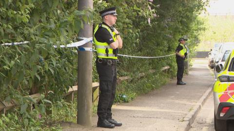 Two police officers standing on a pavement in front of a wooded area that has been cordoned off with blue and white police tape. There is a police car among vehicles parked on the road in front of them.