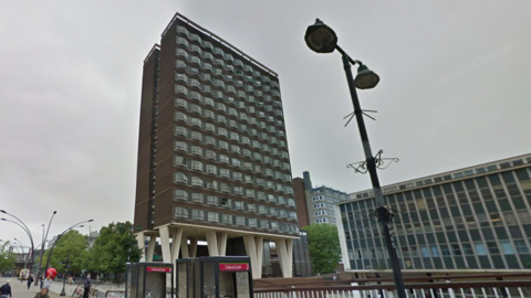 A 14-storey block of flats in Basildon, the building is made of house bricks with V-shaped white supporting piers at ground level. Each floor has 10 windows on the front face. There are several other modern buildings around it, and some old phone boxes in the foreground.