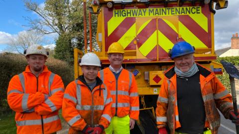 Four highways maintenance staff in high visibility jackets and with hard hats on smile for the camera. In the background is a fluorescent yellow Highways vehicle. 