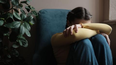 A woman is sitting, hugging her knees with her head buried in her arms. There is a plant beside her.