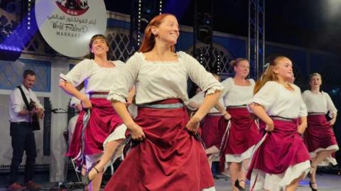 A group of Manx dancers performing on a large stage. They are wearing white square-necked tops and red and white layered skirts. There is a guitarist playing on the stage behind them.
