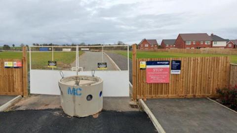 A building site entrance that is blocked by gates and a column of concrete. There are red brick houses in the background to the right and a road behind the gates with grass on either side. 