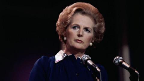 Margaret Thatcher has an auburn bob and large white pearl earrings. She is wearing a dark blue dress with a white collar and is standing in front of two black microphones on stands