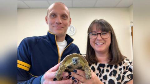 A tortoise looking directly into the camera and looking content and happy with himself. He is being held up by Mr and Mrs Frost. Mr Frost is smiling and wearing a blue fleece and Mrs Frost is also smiling, wears glasses with brown hair, a fringe and a cheetah-print top.