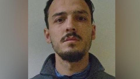 A man wearing a grey fleece with a high collar is looking at the camera. He has dark hair and beard.