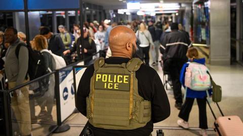 The back of a male ICE agent wearing a vest that reads police and ice below it, in capital letters. He is standing in an airport - behind him are lots of people, some with luggage.