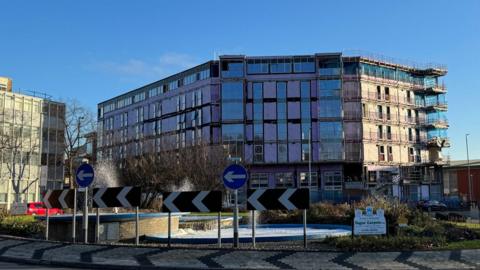 A seven-storey building, which is partially completed, is surrounded by scaffolding. There is a roundabout in the foreground of the image.