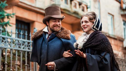 Two people - a man and a woman - dressed in Victorian outfits walking through a street. 