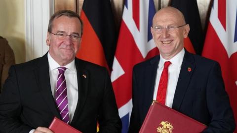 Healey and Pistorius smile, standing in front of the Union Jack and German flags, holding red binders