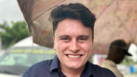 A young man is centre frame and smiling to camera. He has dark hair and is wearing a navy blue shirt.