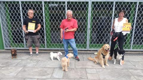 Three dog walkers with their dogs on leads holding Poo Bag Partners posters 