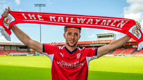 Jordan Gibson is wearing a red and white Crewe Alexandra home shirt and is holding a club scarf above his head with both arms stretched out. The scarf has Crewe's nickname The Railwaymen written on it in white letters. He is standing on the pitch at the Mornflake Stadium in bright sunshine with two stands in the background.