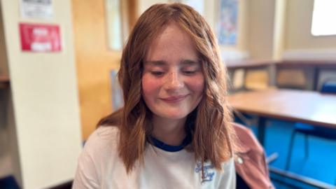 Evie Roberts smiles in a classroom. She is wearing a white t-shirt and has curly auburn hair. Tables and chairs are in the background in what looks like a classroom setting.