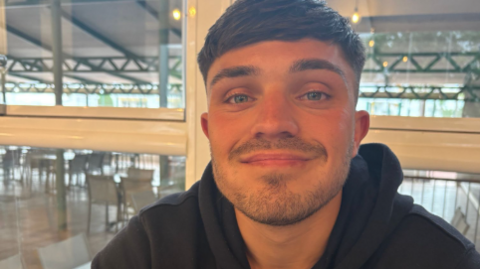 Sam Collard, a young man with dark hair, stubble, and blue eyes. He is wearing a black hoodie.