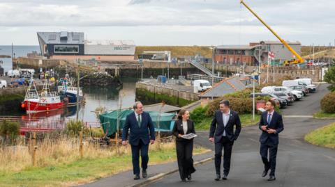 A group of people walk up the hill away from Eyemouth harbour with a busy dock area in the background filled with boats and cars in a car park
