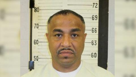 A mugshot of Tony Carruthers. He wears a beige shirt, has a mustache, and stares straight ahead. 