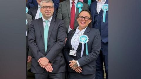 Kathryn Shaw stands next to council leader, Mark Arnull. They both wear suits and Shaw has a pale blue rosette indicating Reform UK on her jacket lapel. Other members of the Reform Party are stood behind them but you cannot see their faces.
