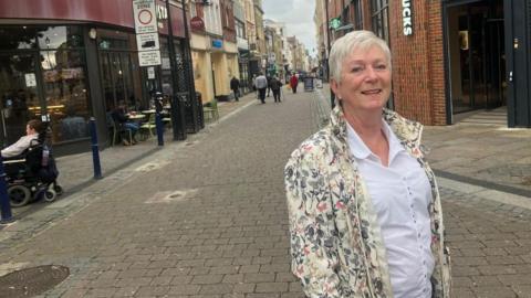 Iris Smith on Gravesend Street is wearing a white blouse and a pale jacket covered in patterns of plants and butterflies. Behind her a group of people are walking down the road