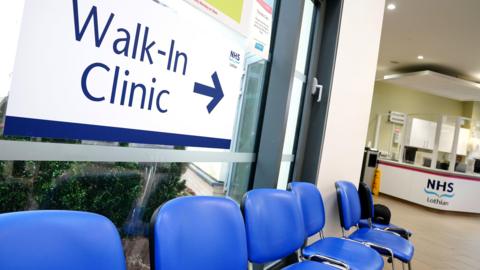 Reception area of the first GP walk-in clinic at Wester Hailes Healthy Living Centre in Edinburgh - six blue chairs are below a sign reading Walk-In Clinic, with an arrow pointing to a reception desk marked with the NHS Lothian logo. 
