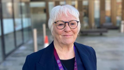 Danielle Stone smiling at camera and wearing a navy blue jacket and glasses standing in front of council offices in Northampton
