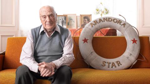 An elderly man in a striped shirt, grey tank top and grey trousers sits on a sofa next to a life buoy ring with the name Arandora Star on it