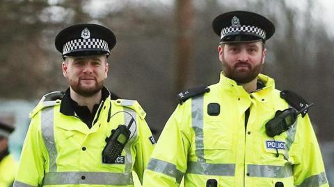 Two bearded police officers walking towards camera in high vis jackets and hats, one of them smiles