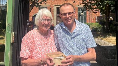 Standing in front of green gates, a tall and slender Filip Krapels wearing a short sleeve blue shirt, hands the cigarette case lid to Diane Owen, an elderly lady with white hair and glasses. Each has one hand supporting the display box in which it was transported