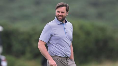 JD Vance looking slightly off camera. He is on a golf course. He is only visible from the waist up. He is wearing a light-coloured polo shirt with a thin horizontal pinstripe design. He is wearing dark beige trousers. He has dark hair and a dark beard. He is holding a golf club in his right hand, which is just out of shot and has his left hand in his pocket.